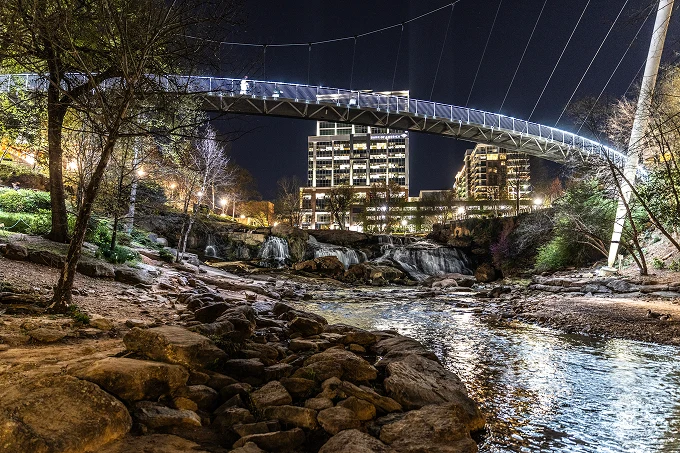 Illuminated suspension bridge over rocky river with buildings and trees at night.