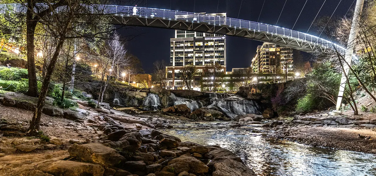 Illuminated pedestrian bridge arching over a rocky river with waterfall and city buildings at night.
