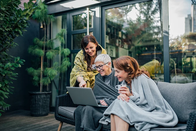 Three women wrapped in blankets sitting on a sofa outdoors looking at a laptop and smiling.