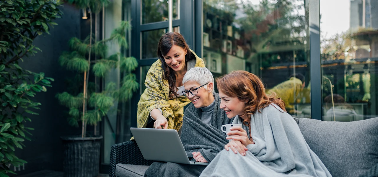 Three women wrapped in blankets smiling and looking at a laptop on a couch outdoors.
