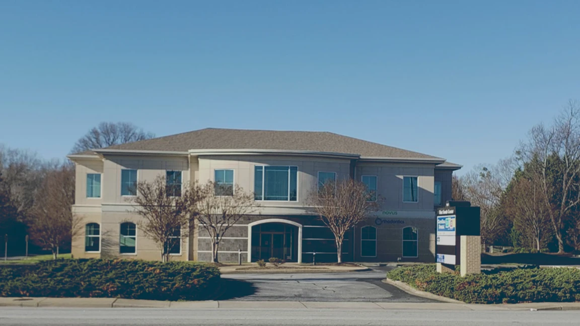 Two-story commercial office building with trees and bushes in front under clear blue sky.