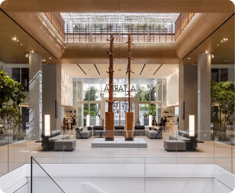 Spacious modern lobby with wooden ceiling, indoor trees, glass railings, and seating areas around two tall wooden sculptures.