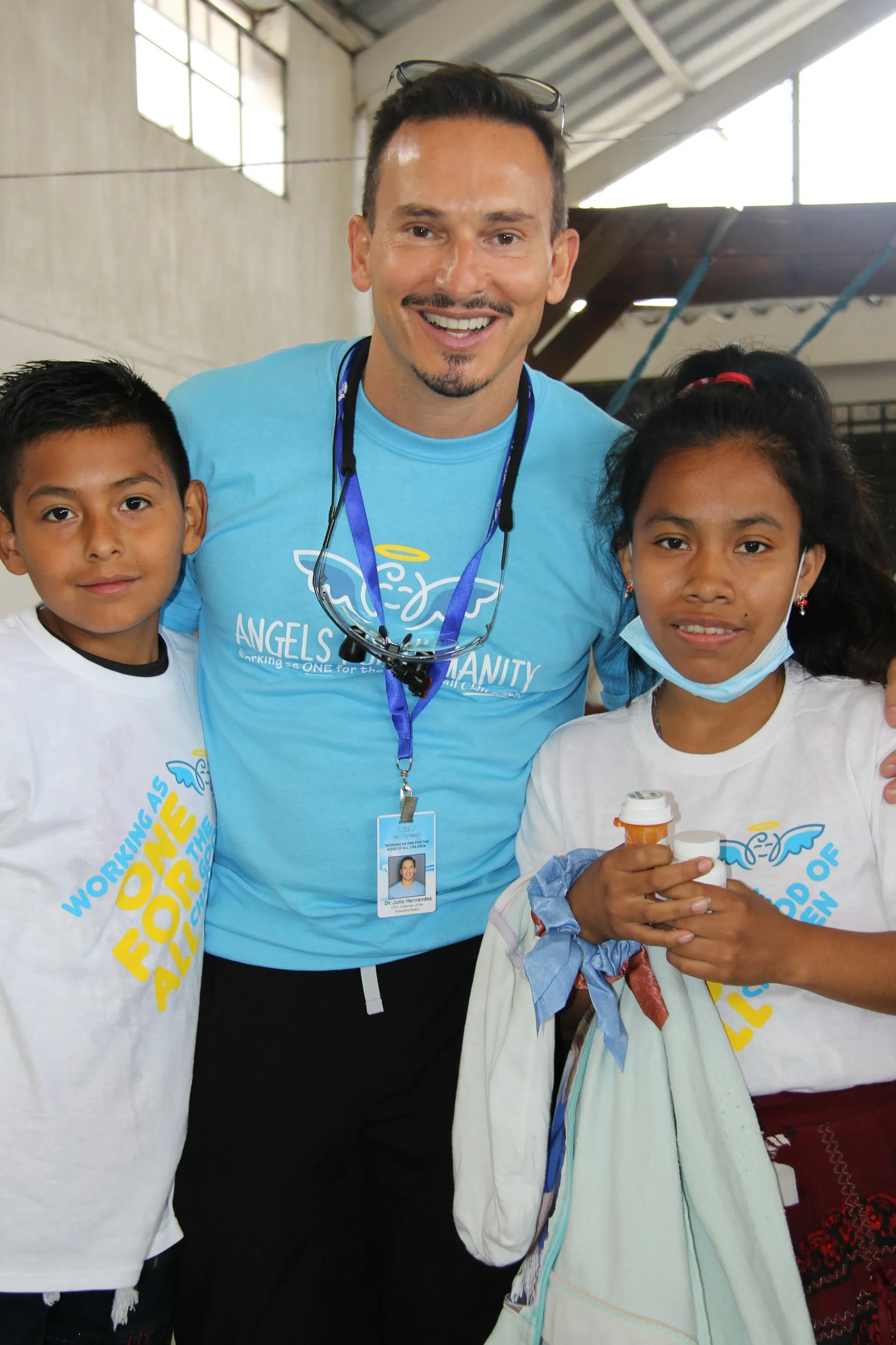 Man in a blue Angels for Humanity shirt stands smiling between two children wearing white t-shirts with the same logo inside a large indoor space.