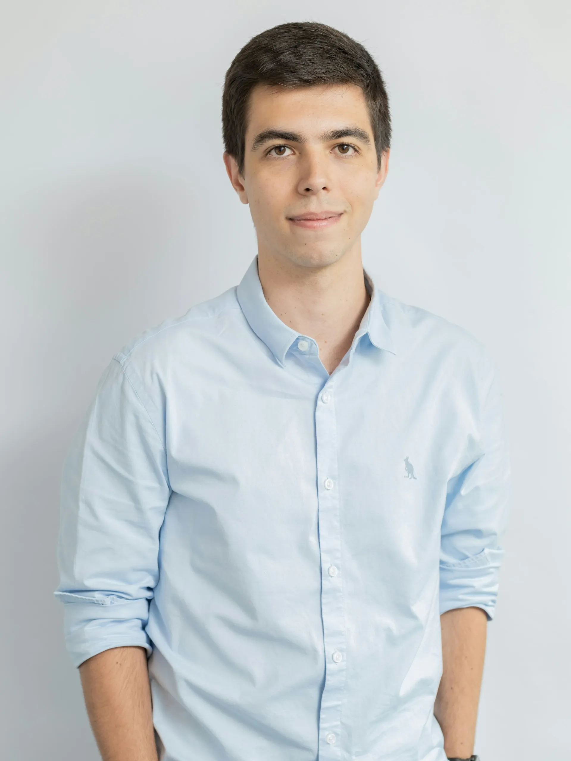 Young man with short dark hair wearing a light blue button-up shirt with rolled-up sleeves standing against a plain light background.