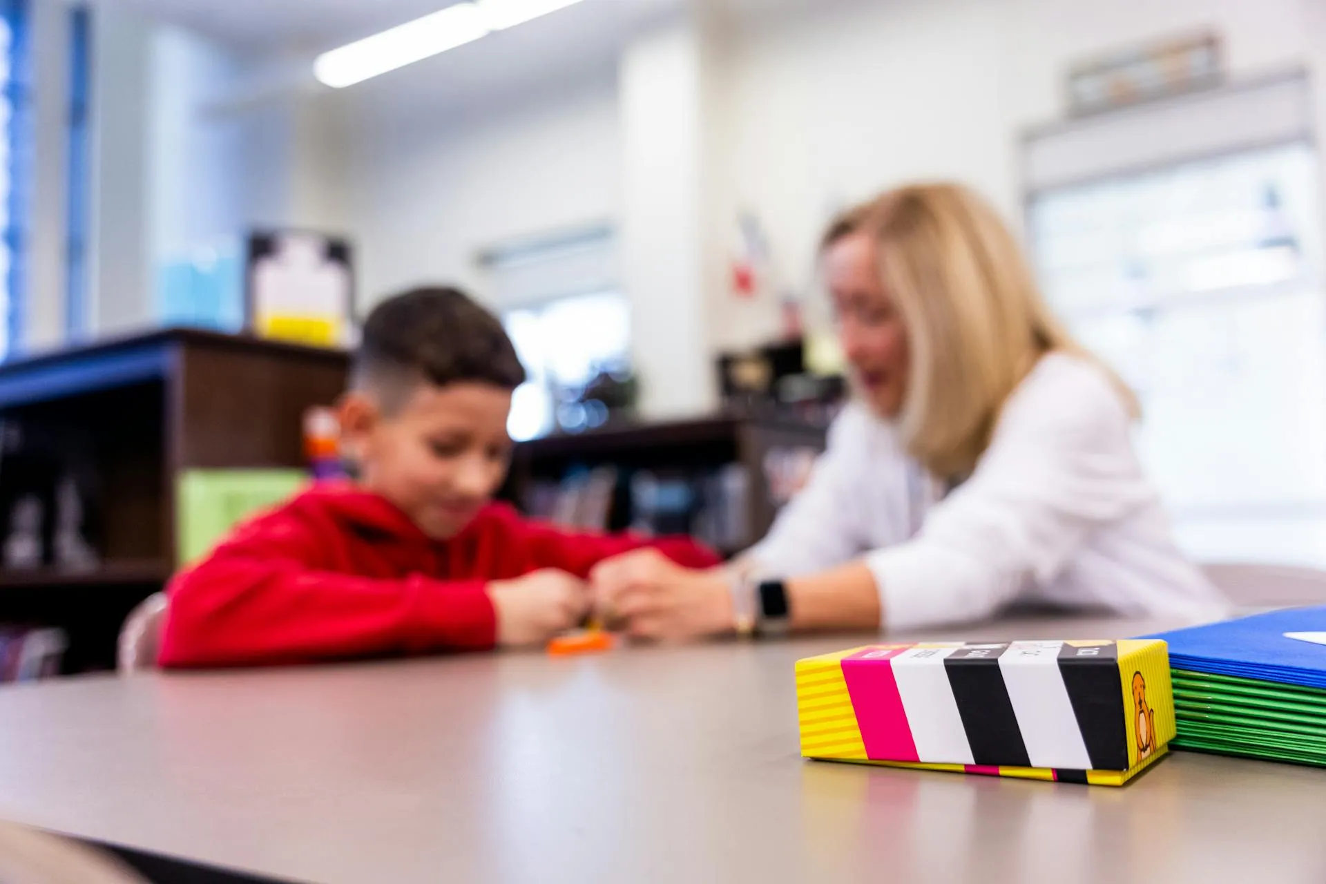 A child and an adult sitting at a table engaged in an activity, with a colorful box and folders in the foreground.