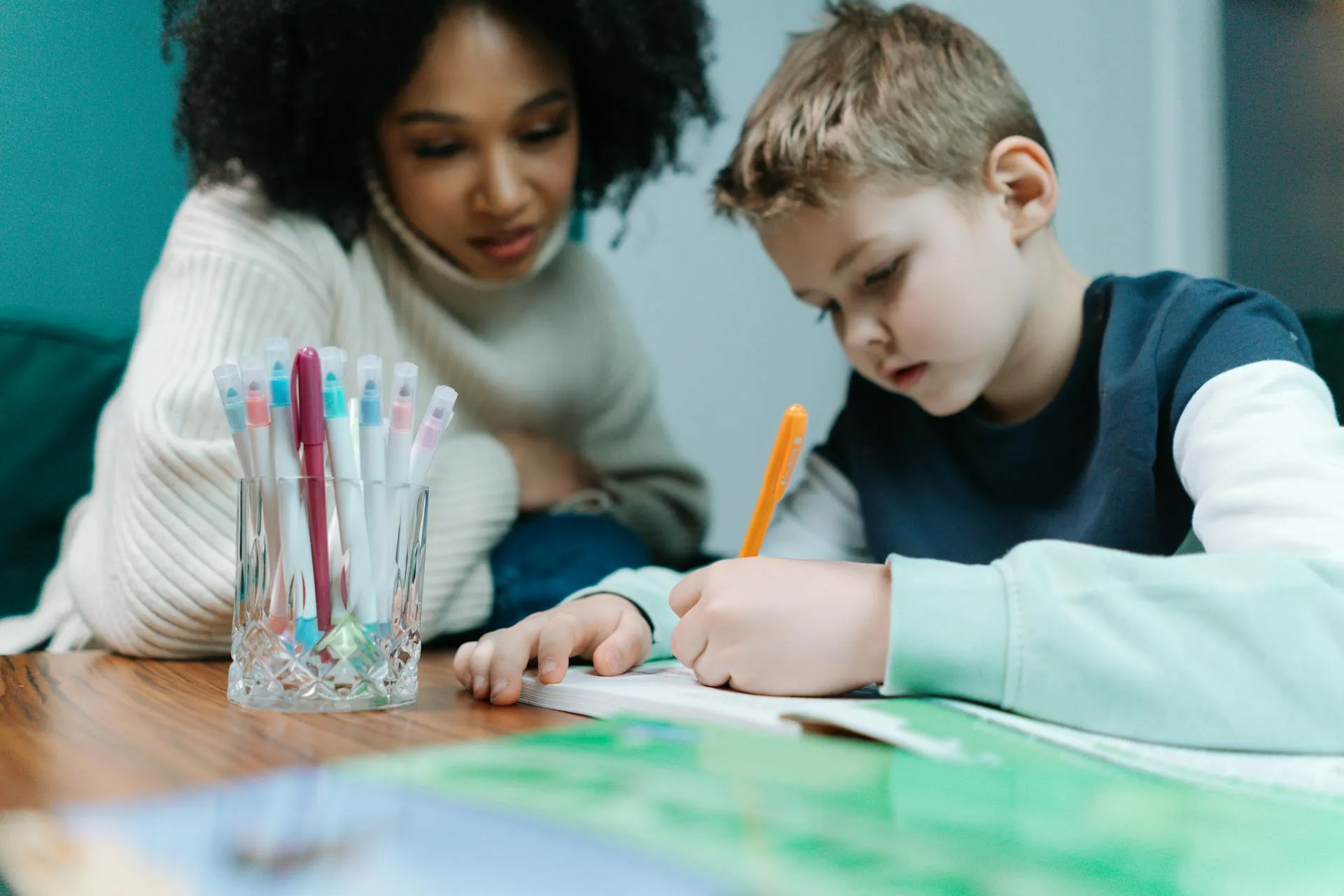 Young boy writing with an orange pen at a table while a woman watches attentively.