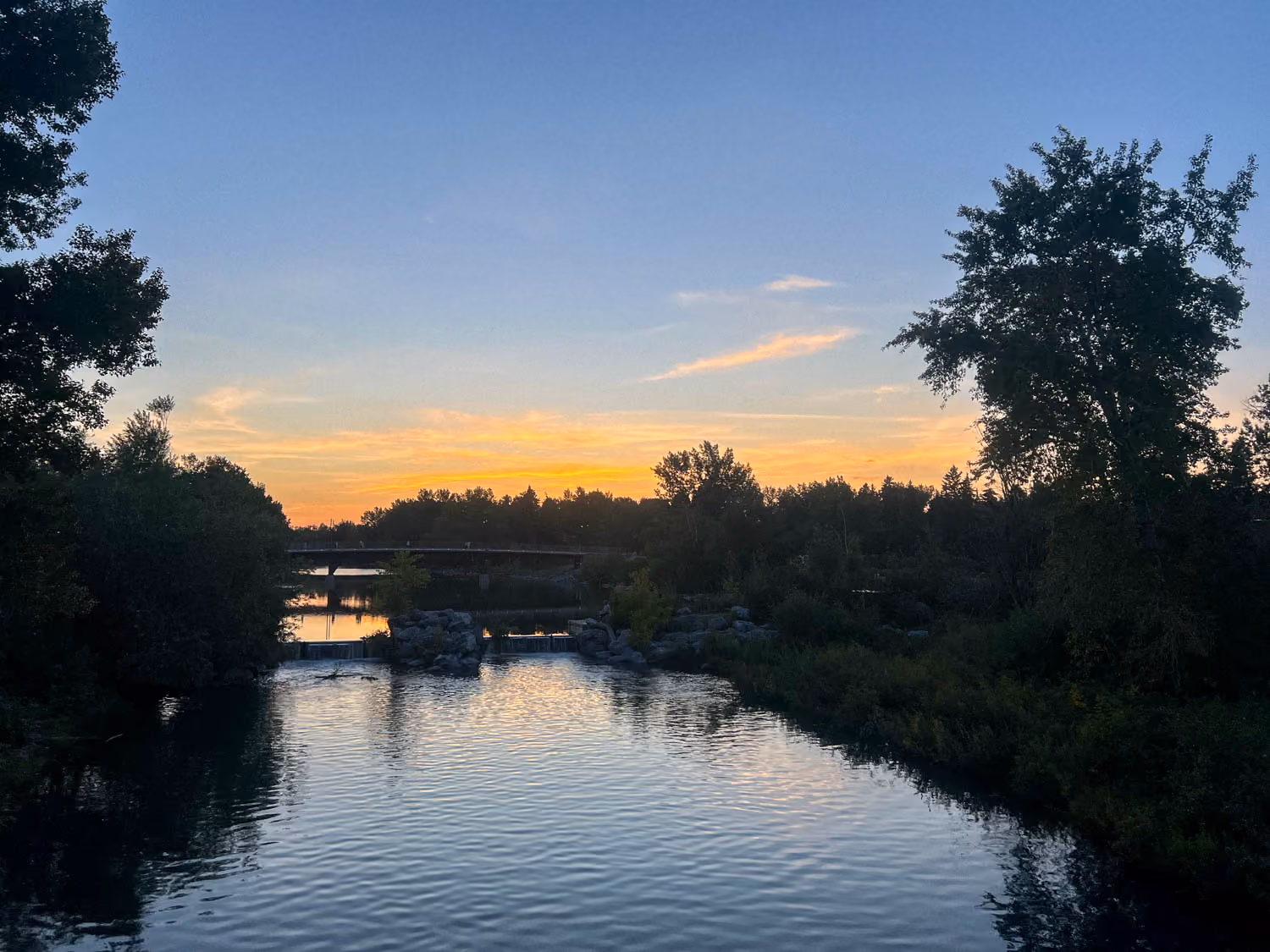 Calm river at sunset with trees along the banks and a small bridge in the distance.