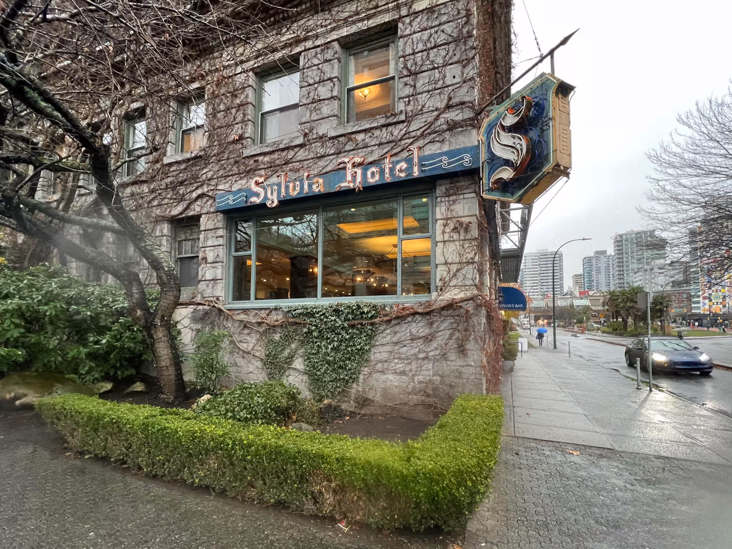Corner of Sylvia Hotel building with ivy-covered walls and a vintage hanging sign in an urban street scene.