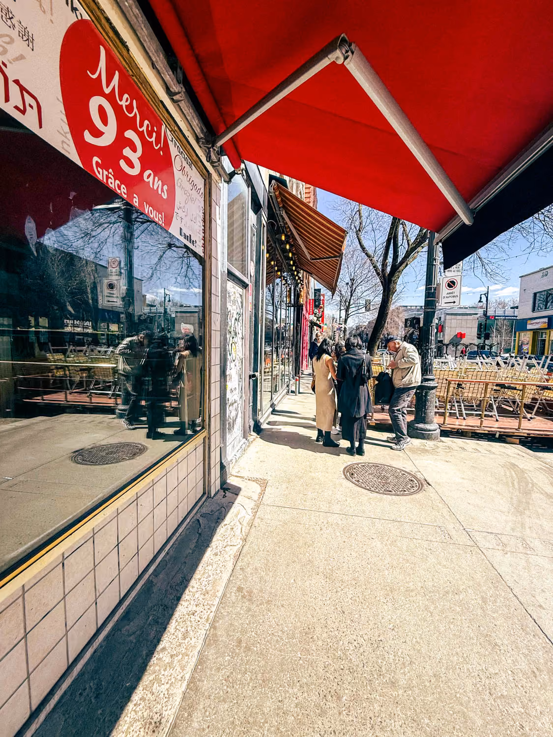 People standing on a sunny urban sidewalk near storefronts with red awnings and outdoor seating.