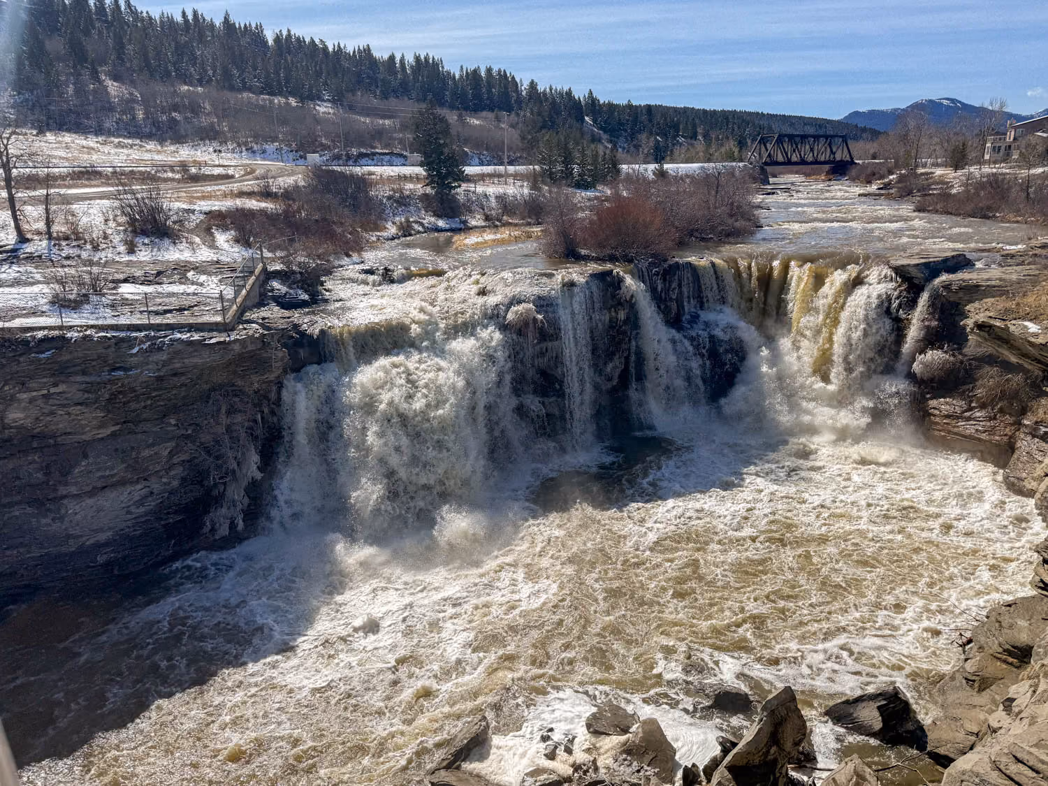 Wide waterfall cascading over rocky cliffs into a turbulent river surrounded by trees and snow patches.