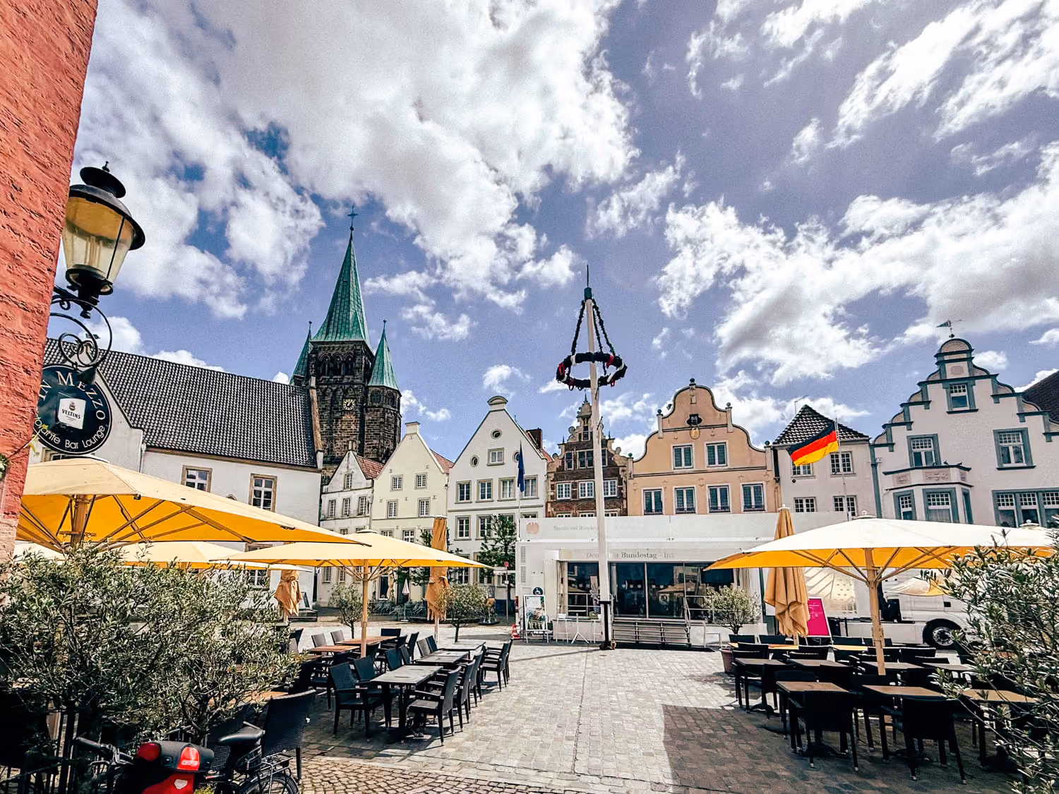 Sunny outdoor café with yellow umbrellas and historic European buildings with pointed church tower.