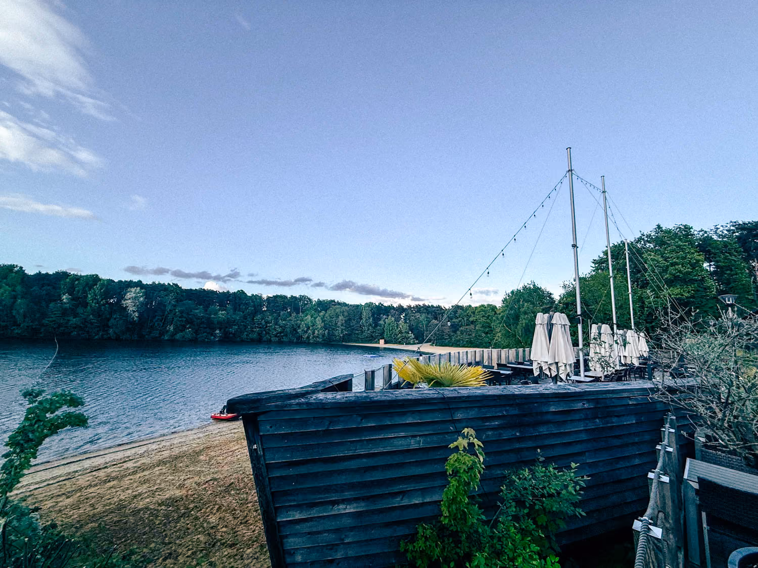 Lakeside view with sandy beach, forest, and a wooden deck with closed umbrellas and string lights.