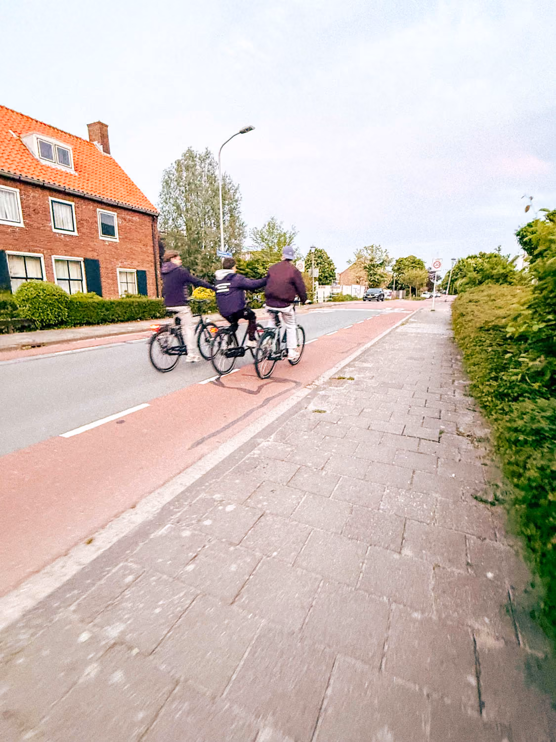 Three people riding bicycles on a street bike lane beside a red-roofed house and greenery.