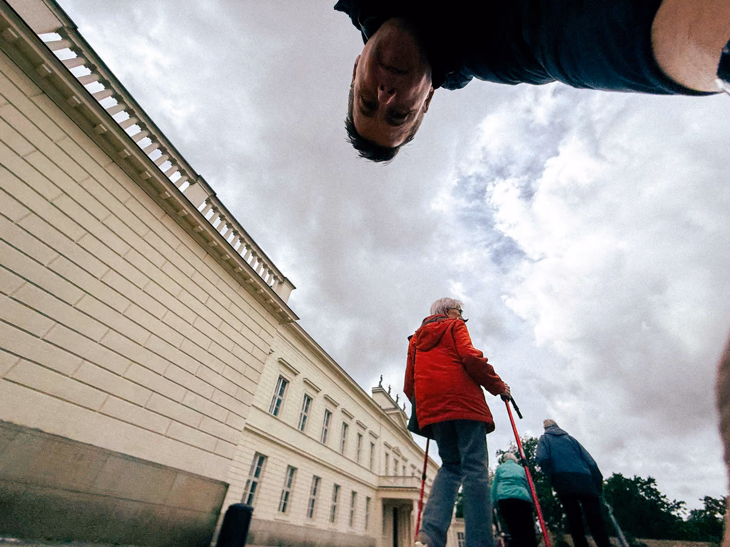 Low-angle view of three elderly people walking with canes near a large cream building under cloudy sky.