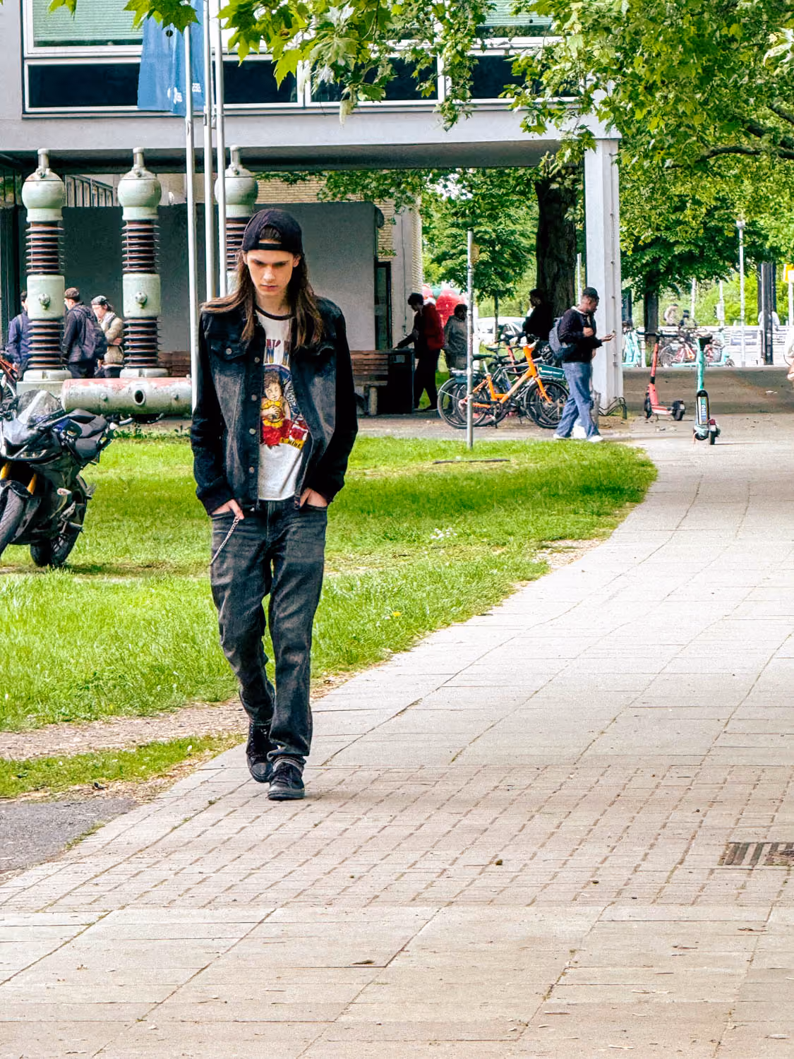 Young man with long hair, black cap, and jacket walks on a city sidewalk near grass and bikes.