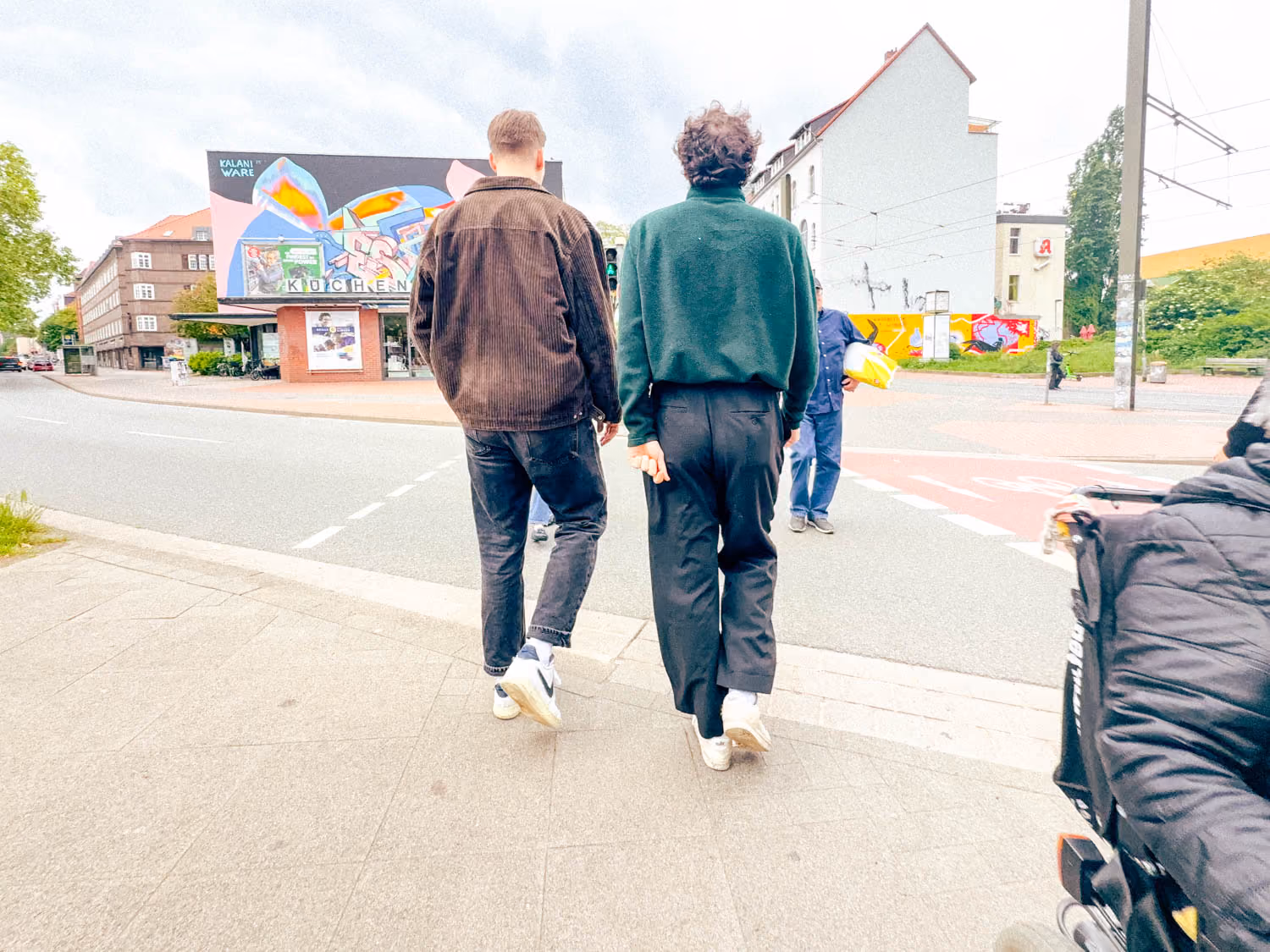 Two men walk on a sidewalk near a street with a colorful mural on a building in the background.