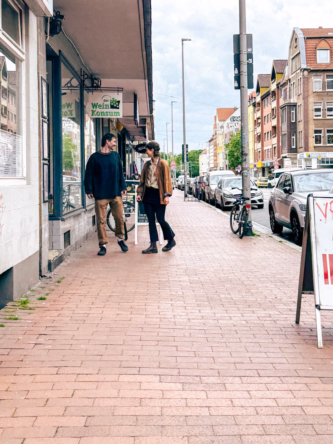 Two people walking and talking on a city sidewalk near parked cars and a storefront sign reading der Wein Konsum.