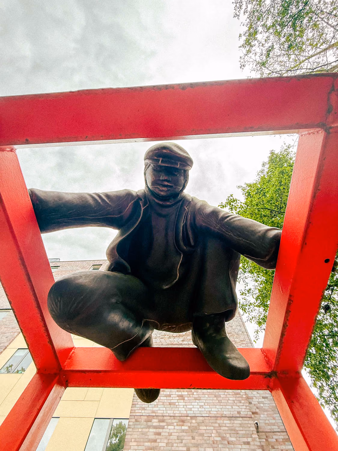 Bronze statue of a crouching man framed by a red metal square viewed from below against sky and buildings.