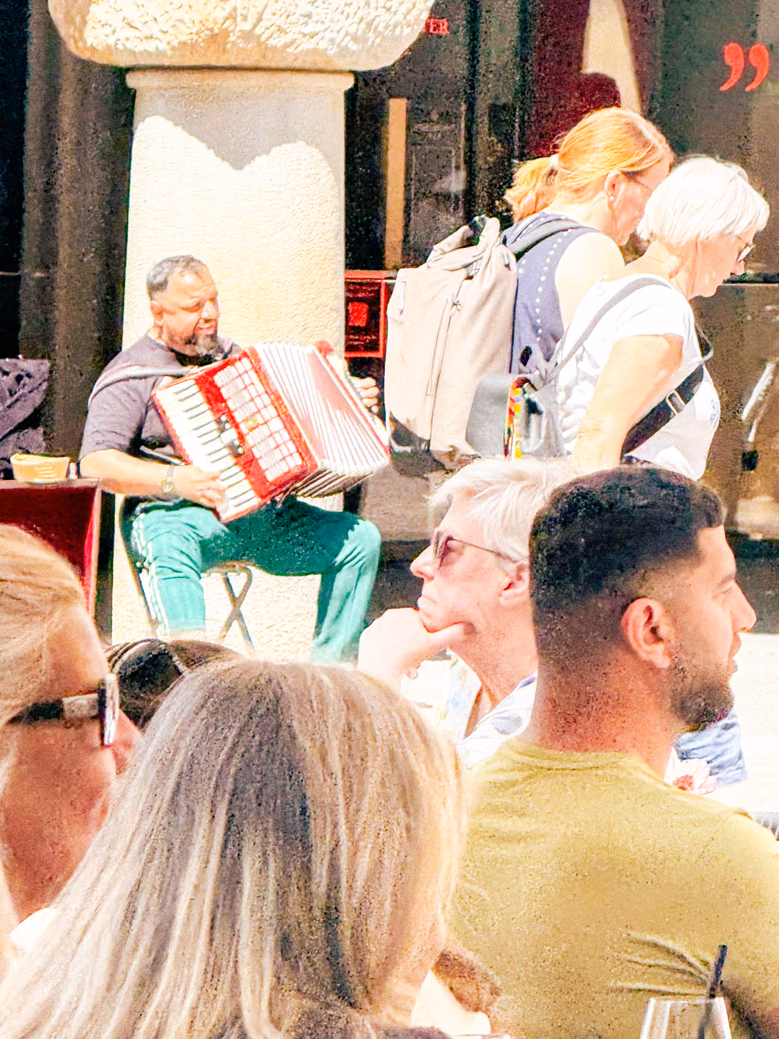 Man sitting on a stool playing a red accordion near a column in an outdoor setting with several people nearby.