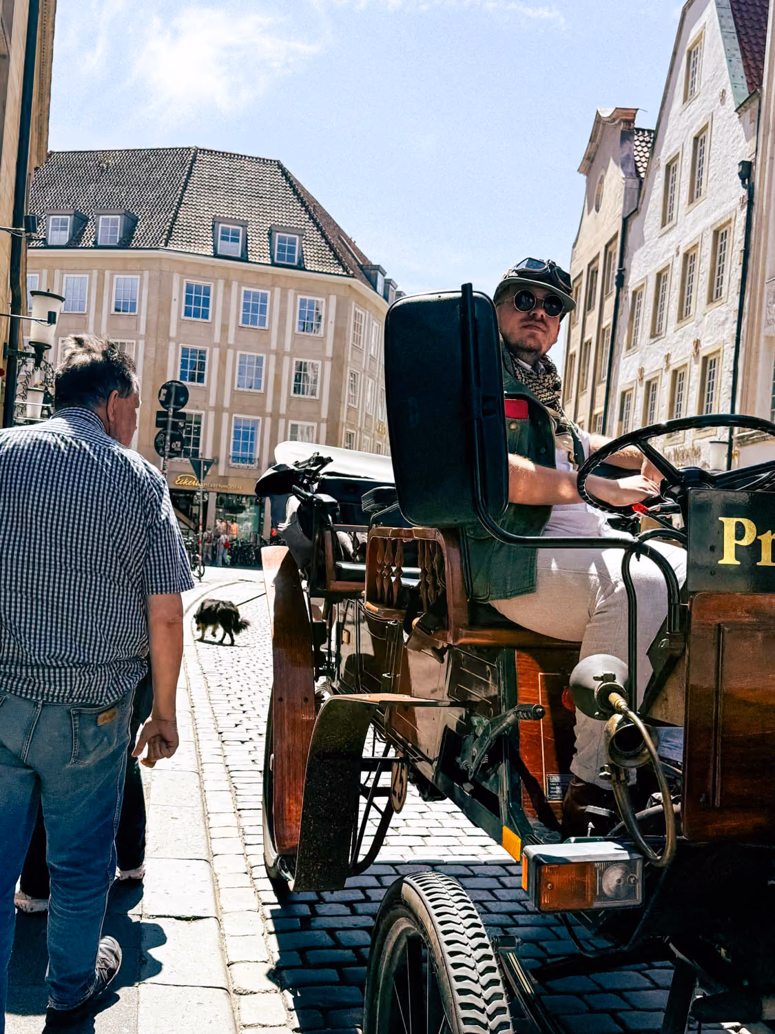 Man wearing sunglasses drives a horse-drawn carriage on a cobblestone street with a pedestrian nearby.