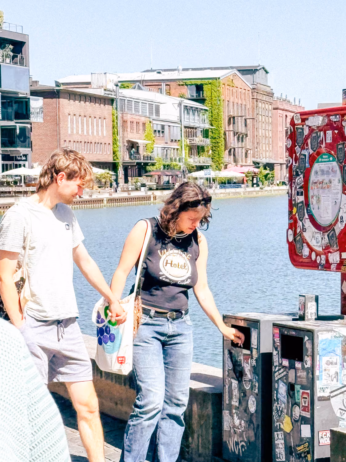 Young couple holding hands near waterfront with buildings and stickers on machines in background.