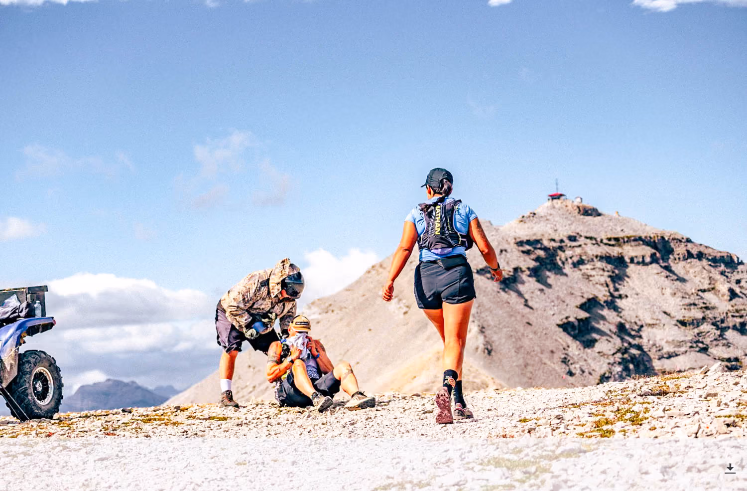 Runner walking toward mountain peak as another rests on rocky trail with helper beside them.