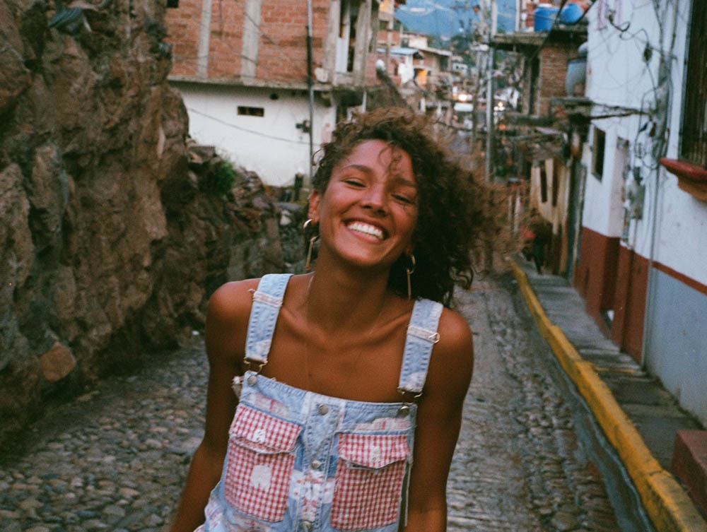 Smiling woman with curly hair wearing denim overalls on a cobblestone street in a village.
