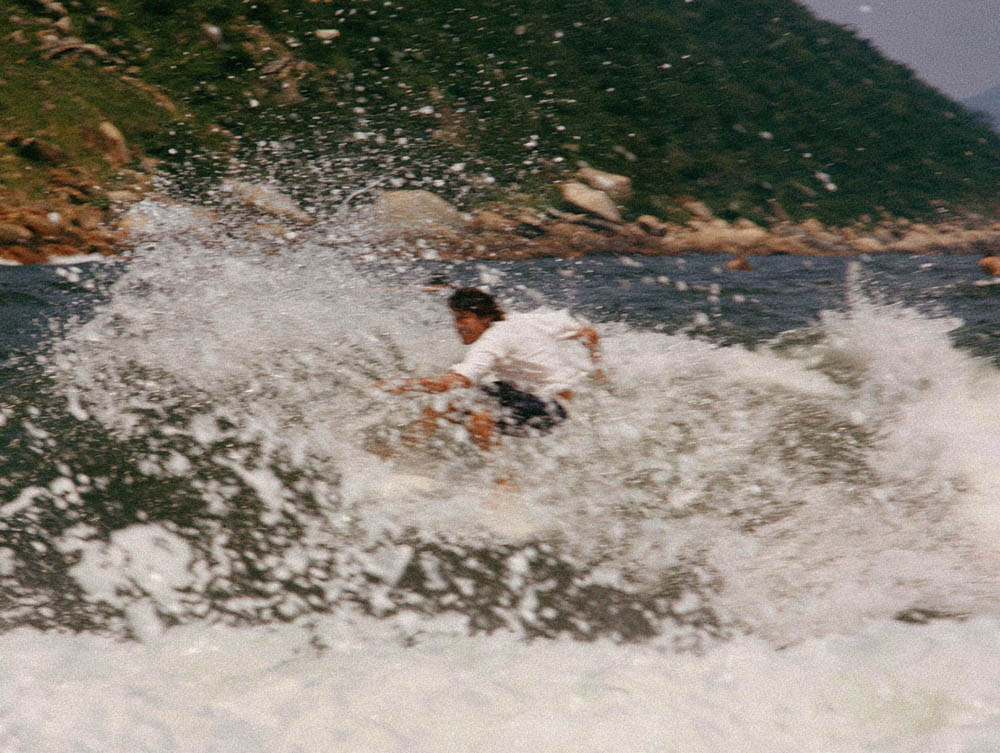 Person surfing on a wave near a rocky shoreline with green hills in the background.