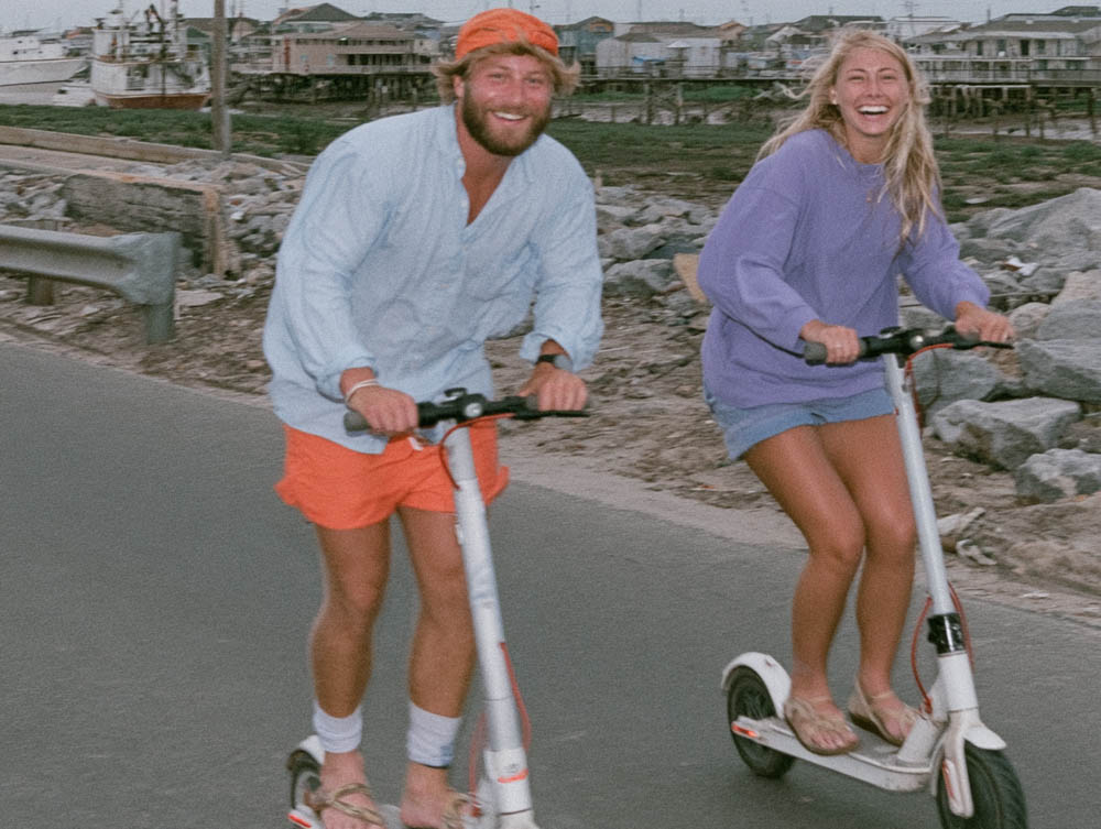 Smiling man and woman riding white electric scooters on a paved road near rocky shore and buildings.