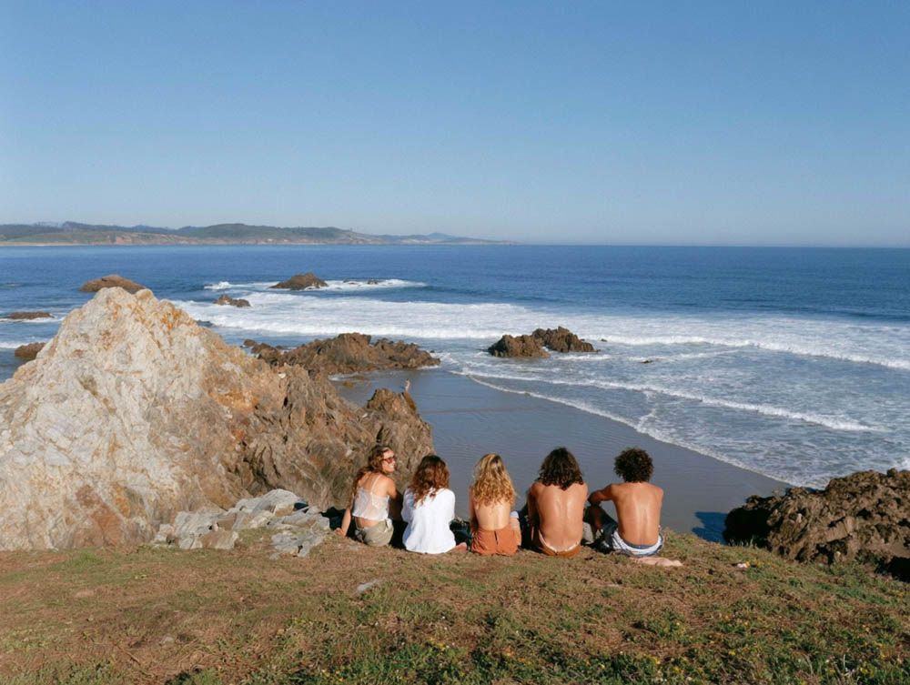 Five people sitting on a grassy cliff overlooking the rocky shore and ocean under a clear blue sky.