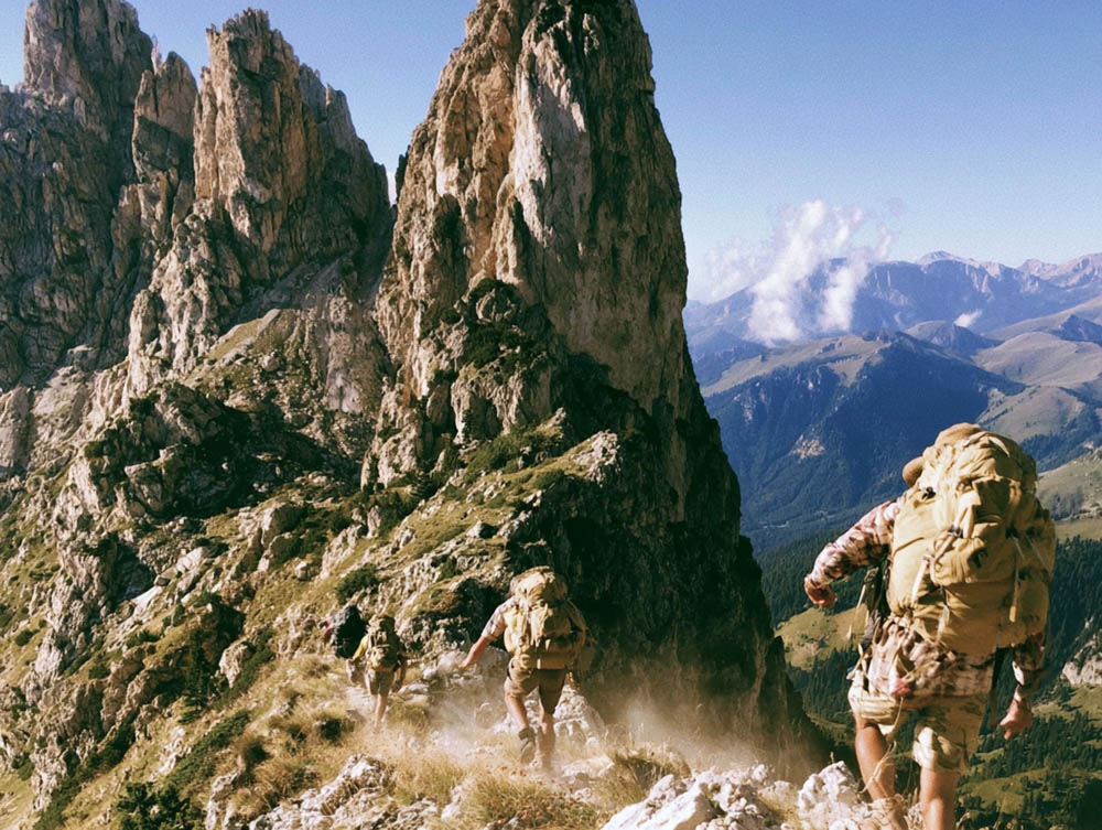 Three hikers with large backpacks trekking down a rocky mountain ridge under a clear blue sky.