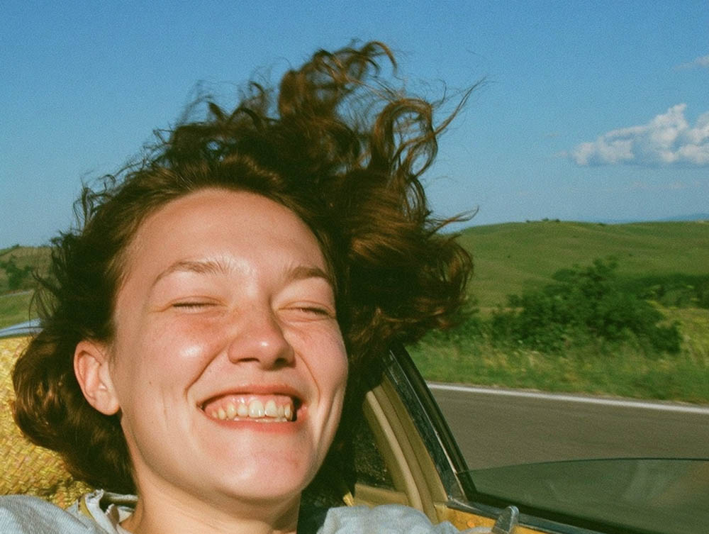 Young woman smiling with eyes closed, hair blowing in wind through open car window on a sunny day.