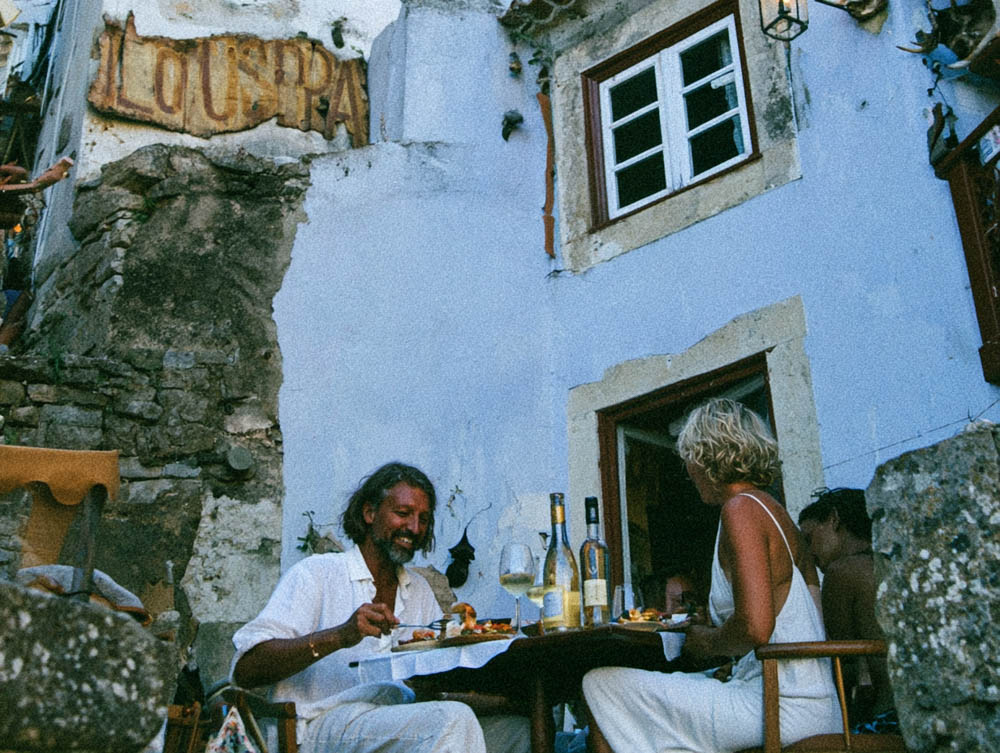 Two people dining outdoors at a rustic stone building with wine bottles and glasses on the table.