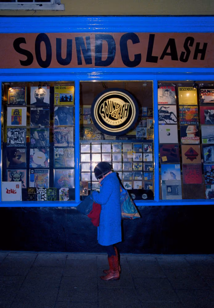 Person in blue coat and red boots standing outside Soundclash record store at night.
