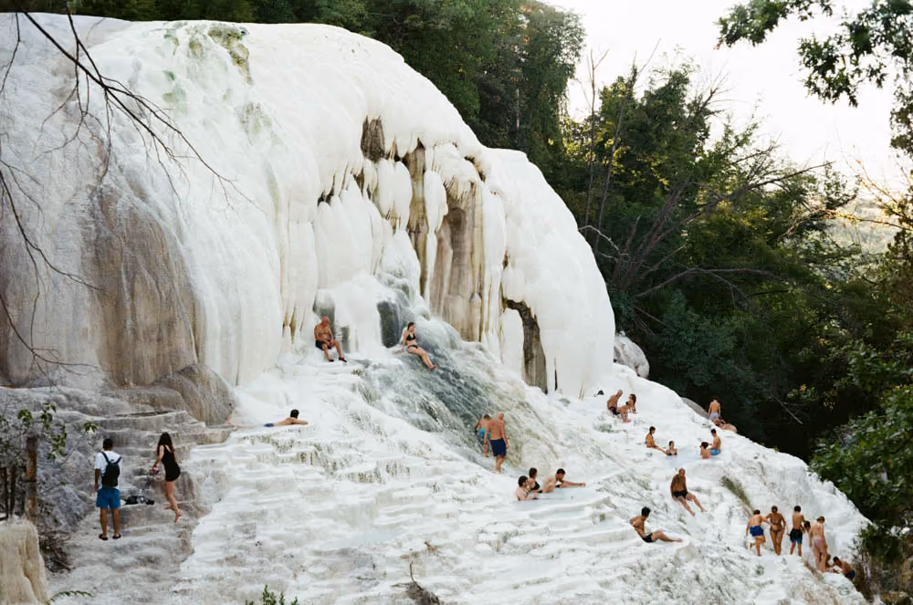 People bathing and sitting on natural white limestone terraces with flowing water surrounded by greenery.