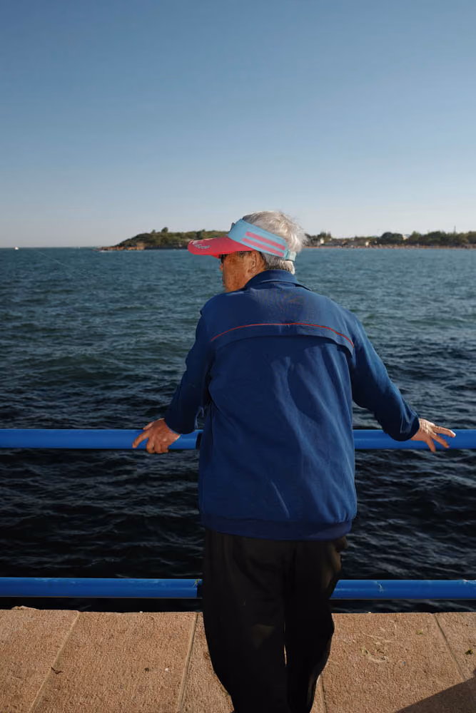Older man wearing a visor and blue jacket leaning on railing by the water with an island in the distance.