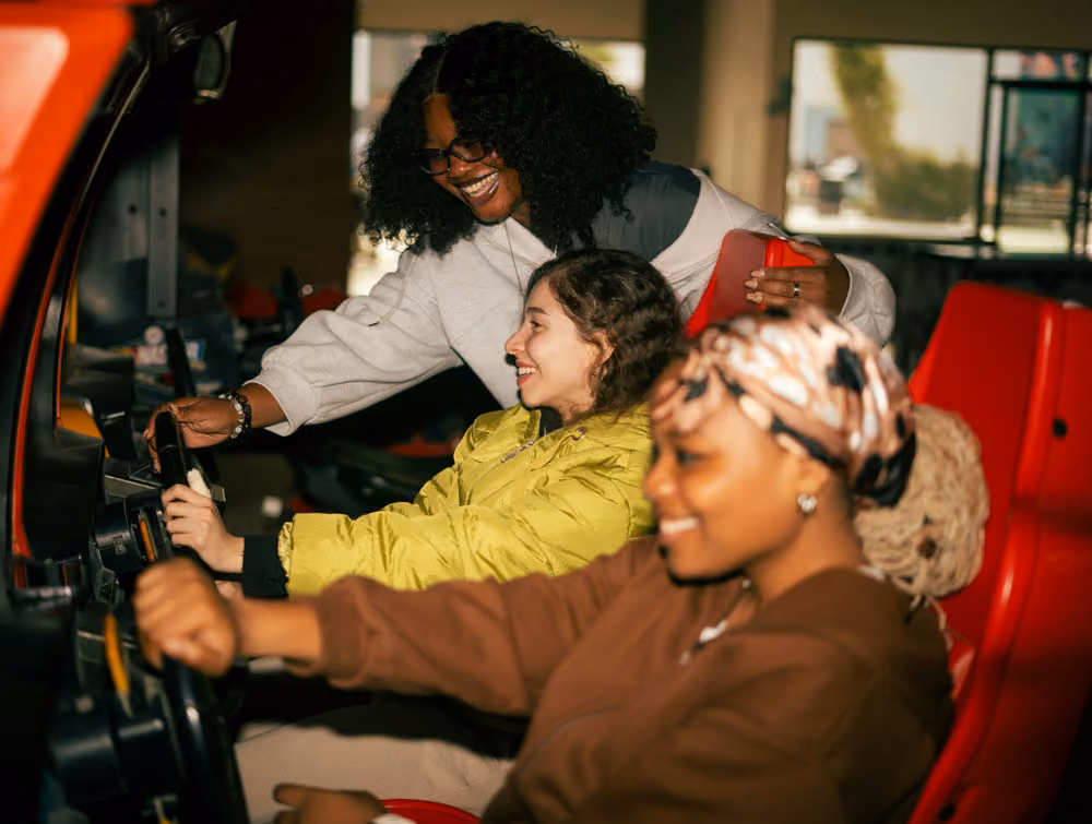 Three young women smiling while playing an arcade driving game seated in red chairs.