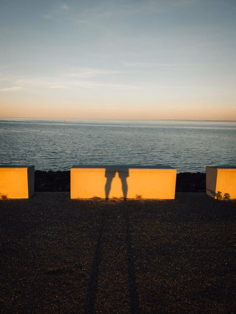 Shadows of two people standing close projected on a concrete wall by a waterfront at sunset.