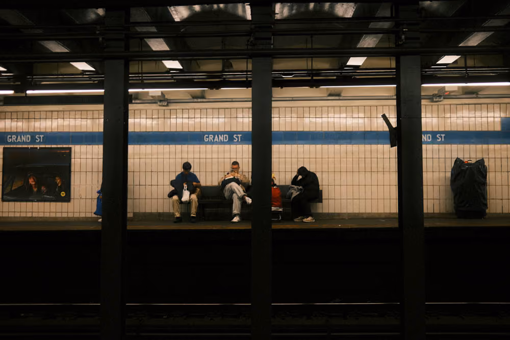 Three people sitting on benches on a subway platform at Grand St station, separated by pillars.