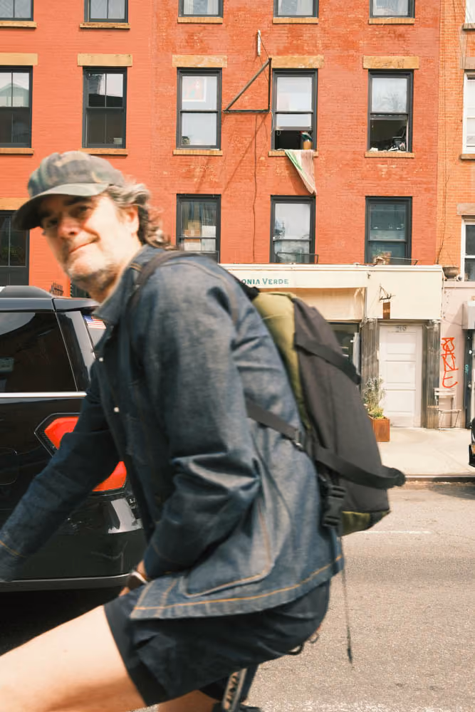 Man wearing cap and denim jacket rides a bicycle on a city street with red brick buildings in background.