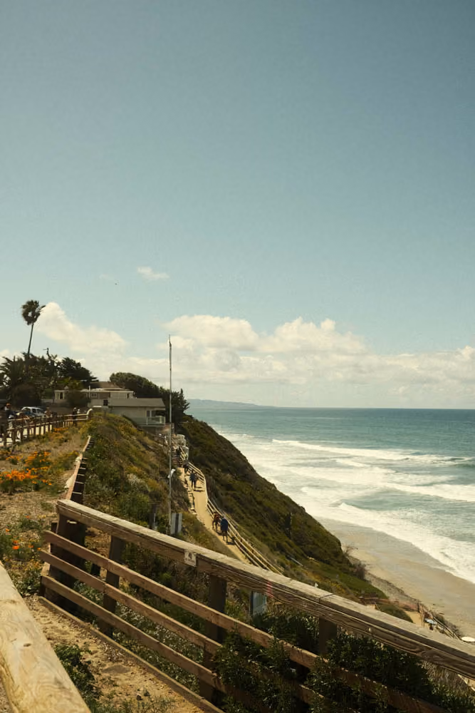 Coastal trail with wooden fences overlooking ocean waves and sandy beach on a sunny day.