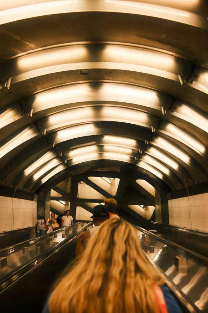 People on an escalator under a modern curved ceiling with bright lights in a subway station.