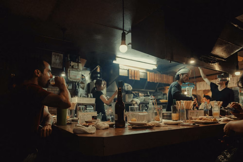 Dimly lit bar with people drinking, a bartender, and various bottles and glasses on the counter.