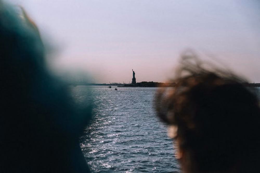 View of Statue of Liberty across water with silhouettes of two people in foreground.