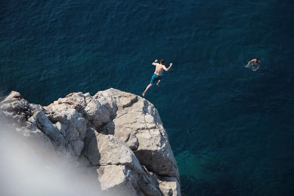 Man jumping from a rocky cliff into deep blue ocean water while another swims nearby.