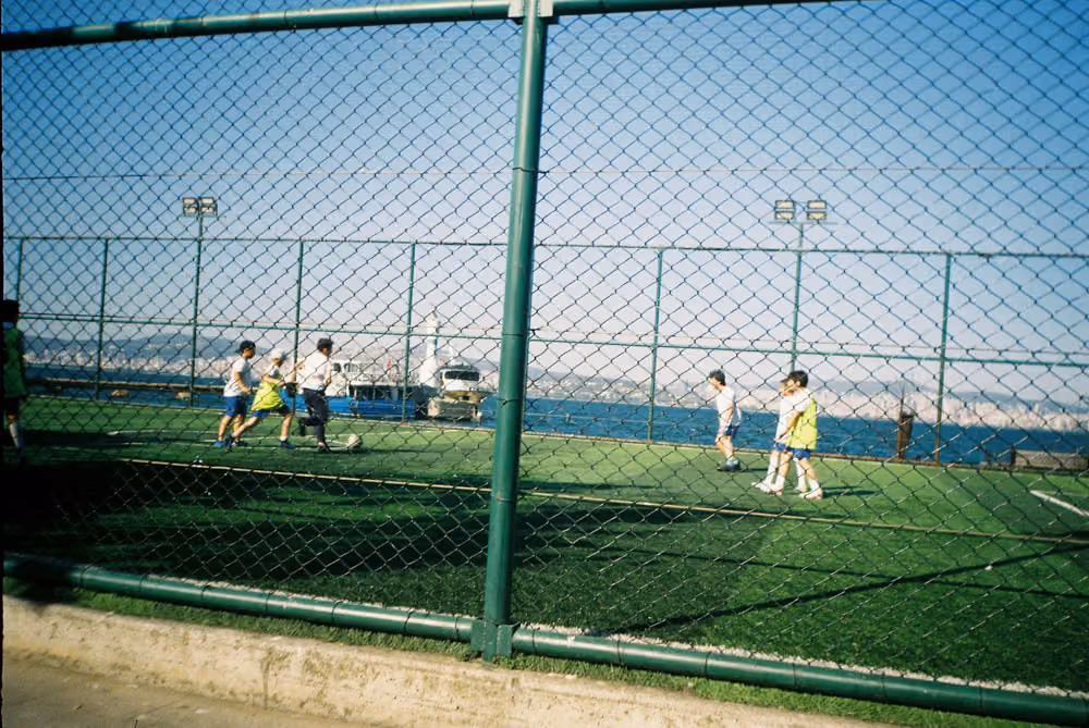 Children playing soccer on a fenced-in artificial turf field near a waterfront.