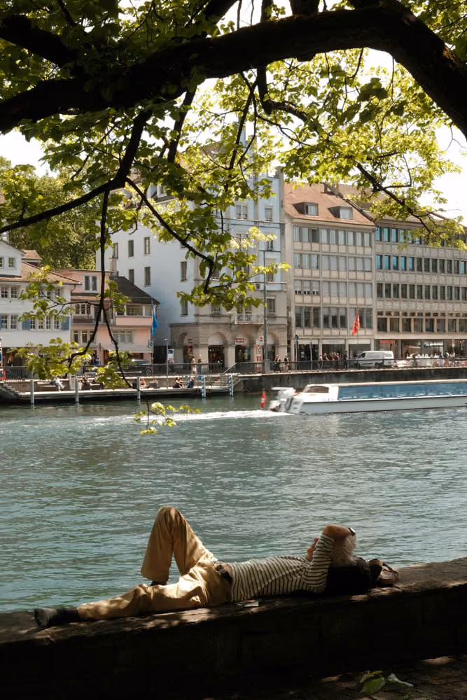 Person lying on a riverbank wall under tree branches with city buildings and a boat in the background.