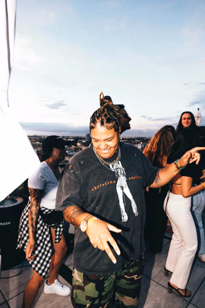 Person with braided hair dancing outdoors with others at sunset on a rooftop.