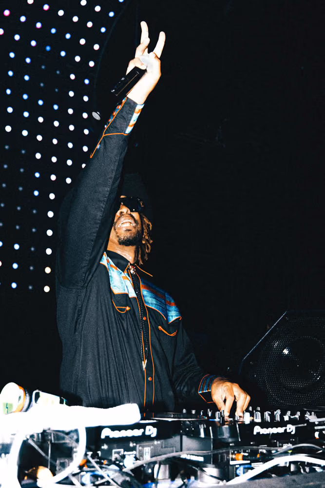 DJ wearing sunglasses and a hat raises a microphone while mixing music on Pioneer DJ equipment at a club.