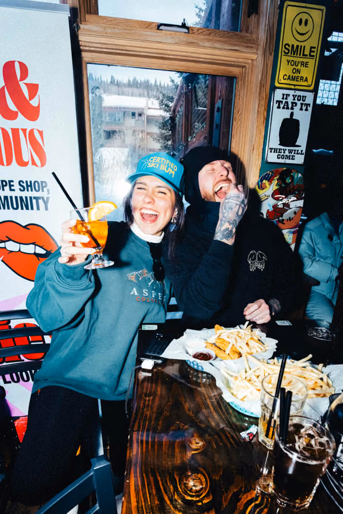 Two people laughing inside a restaurant with drinks and plates of fries on the wooden table.
